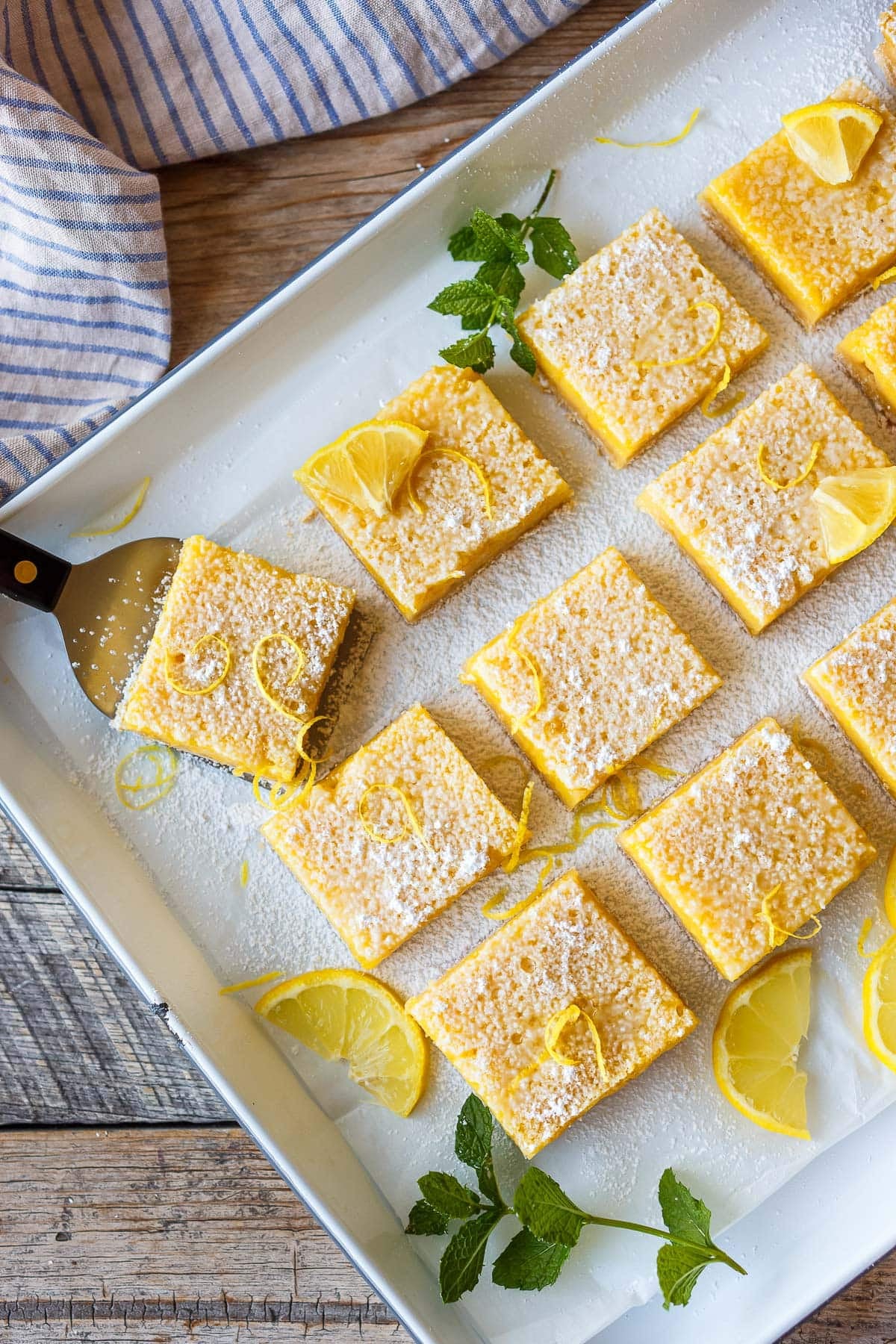 baking tray with clean cut squares of lemon bars topped with powdered sugar and lemon zest and lemon wedges with a spatula picking up a piece.