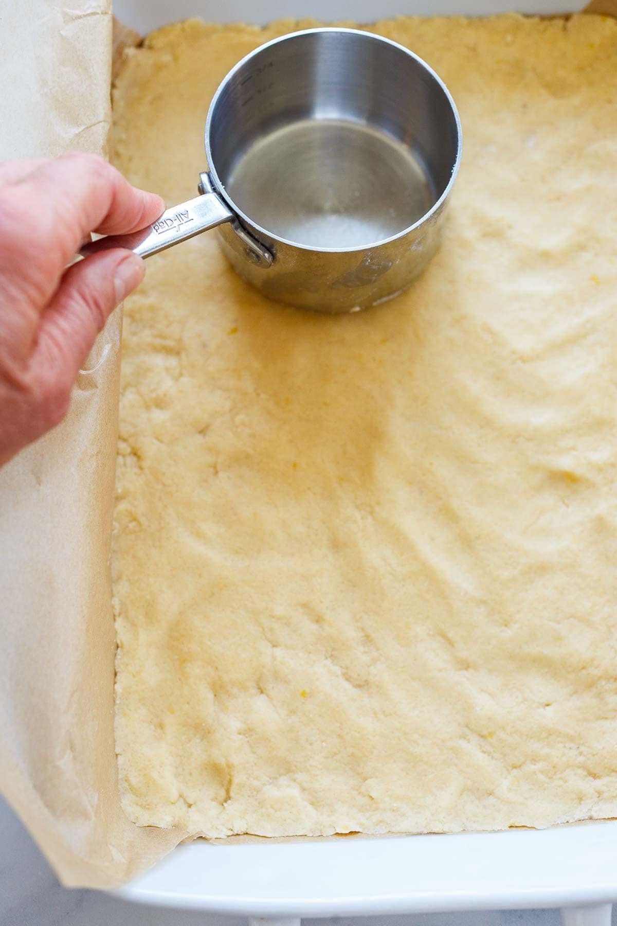 hand pressing the bottom of a measuring cup onto shortbread crust to prepare lemon bars.