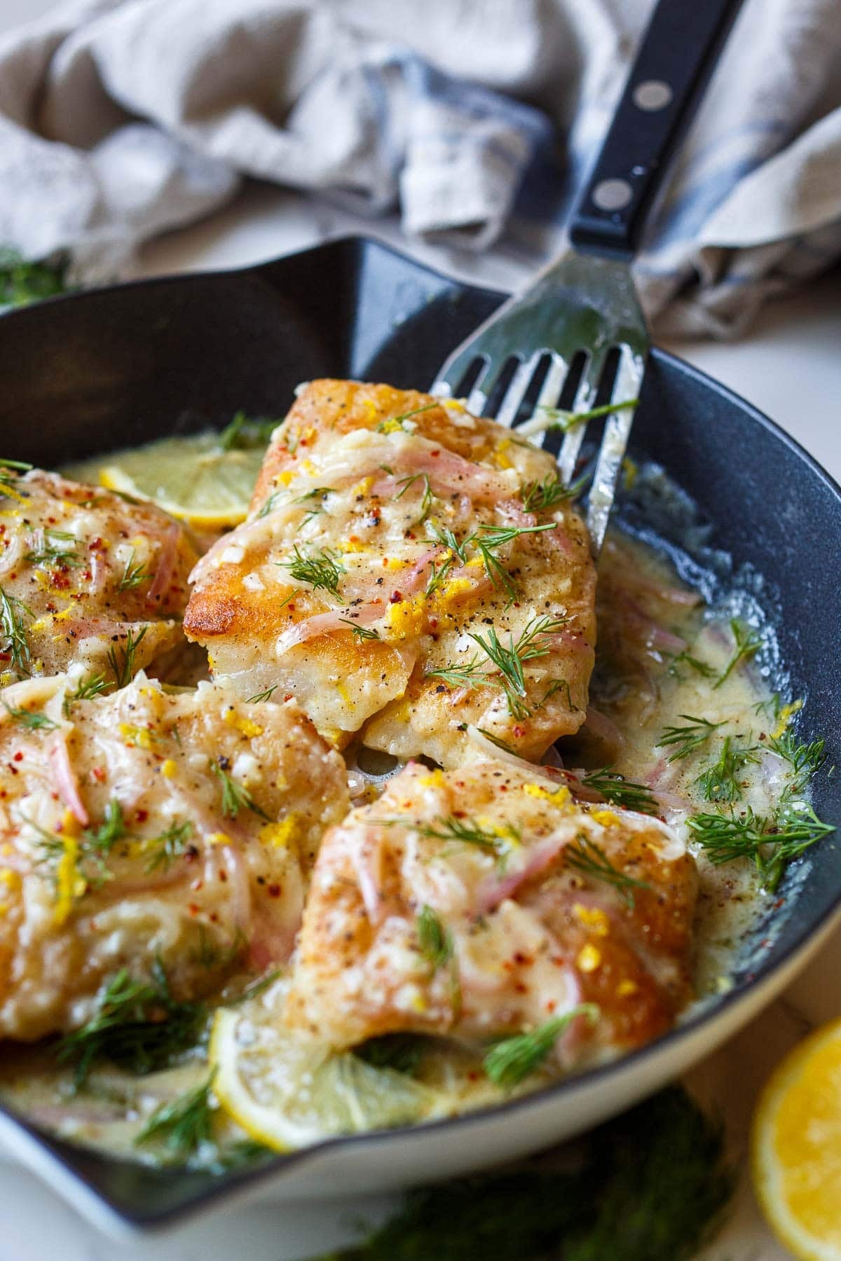 lemon butter cod being lifted from the skillet with a fish spatula, garnished with lemon zest, pepper, chili flakes, and fresh dill.