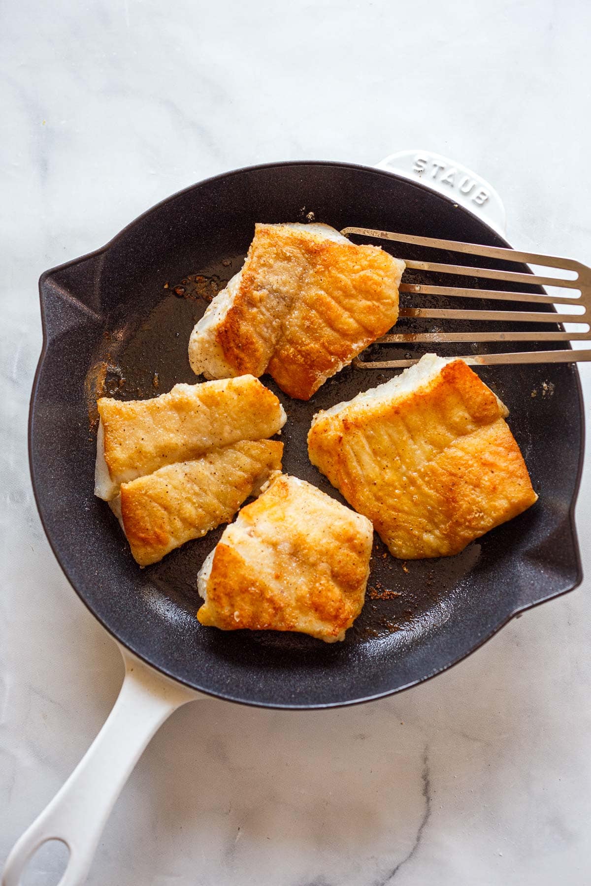 four cod fillets searing in a skillet with a fish spatula.