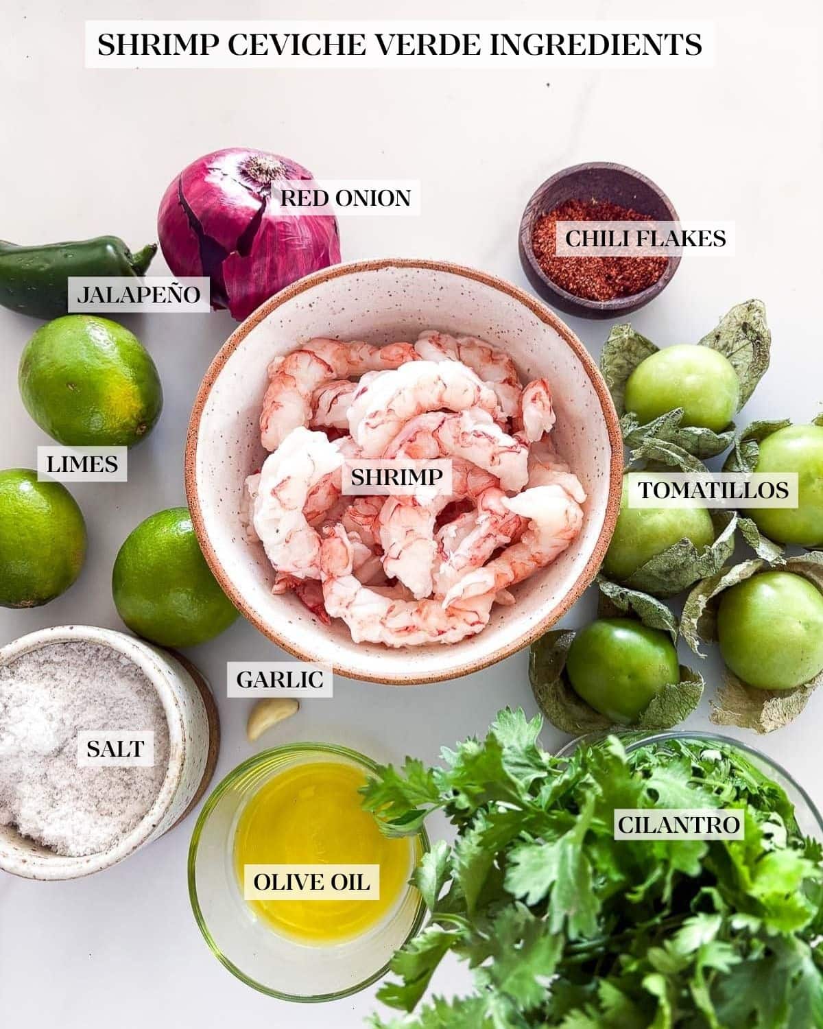 ingredients laid out for shrimp ceviche verde - jalapeño, red onion, limes, pinch bowl with chili flakes, tomatillos, bowl of shrimp, dish of salt, garlic clove, glass bowl of olive oil, and a cilantro bunch.