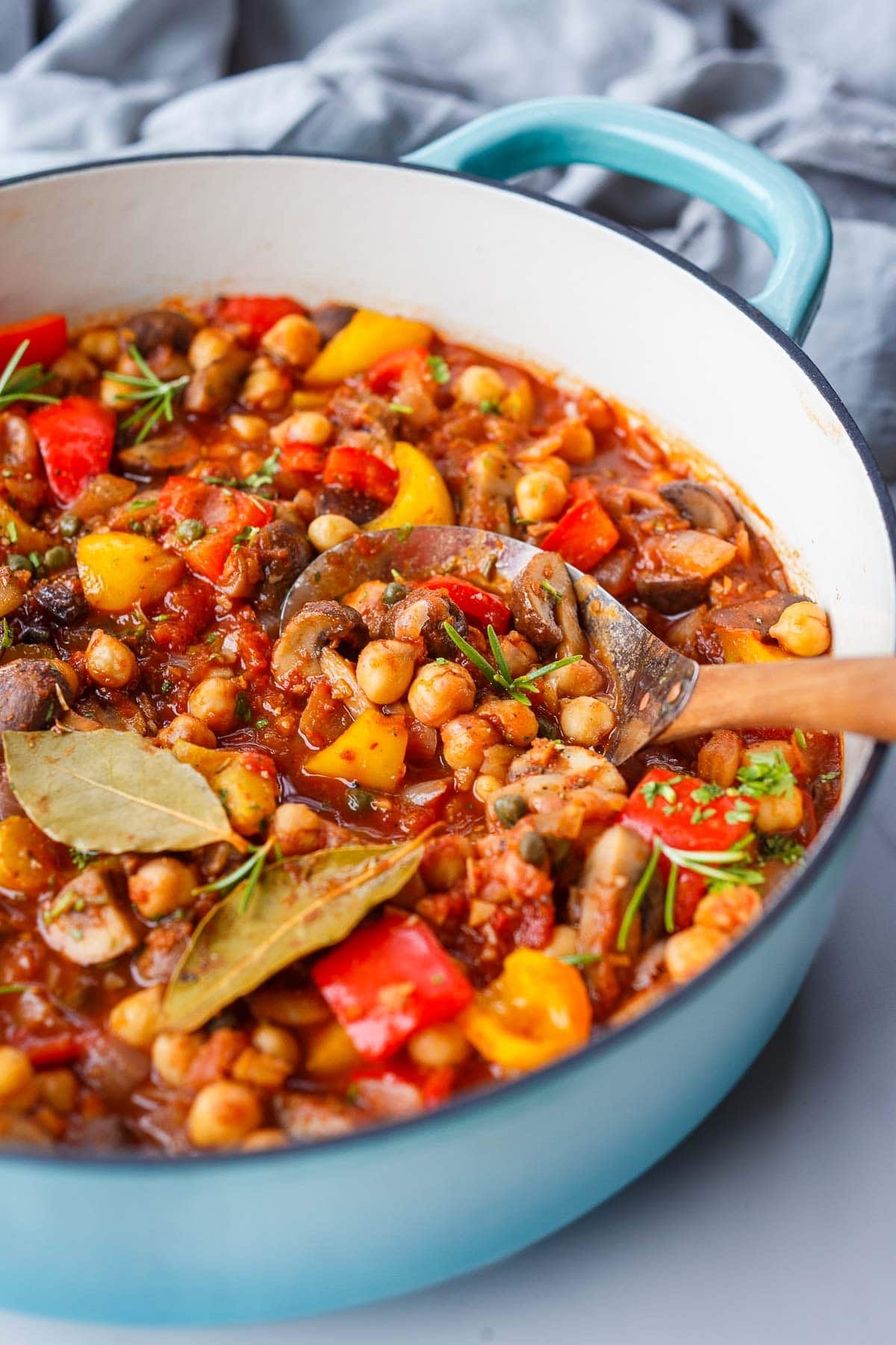 chickpea cacciatore in a dutch oven with mushrooms, bell peppers, rosemary, bay leaves, and capers, with a wood and metal serving spoon.