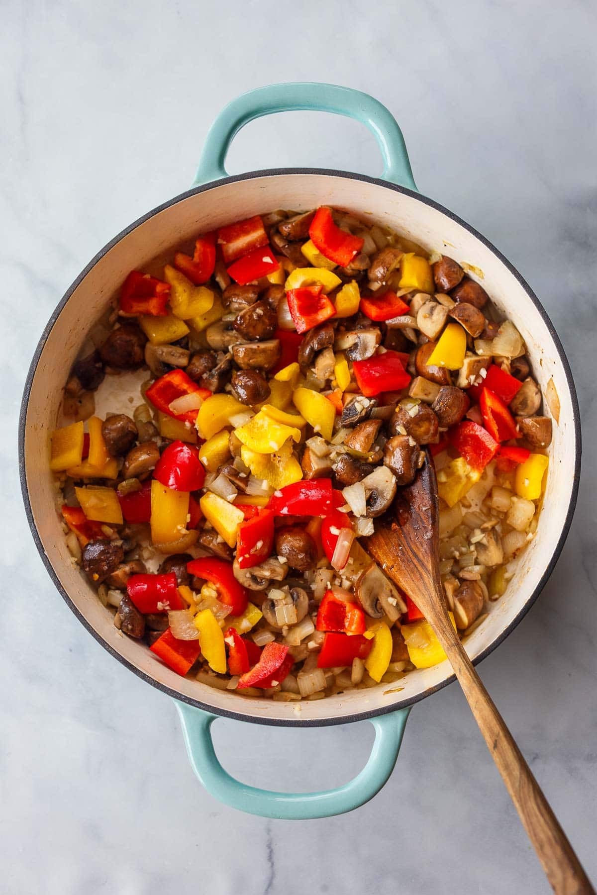 dutch oven with sautéed onions, mushrooms, red bell peppers, and yellow bell peppers with a wooden utensil to stir.