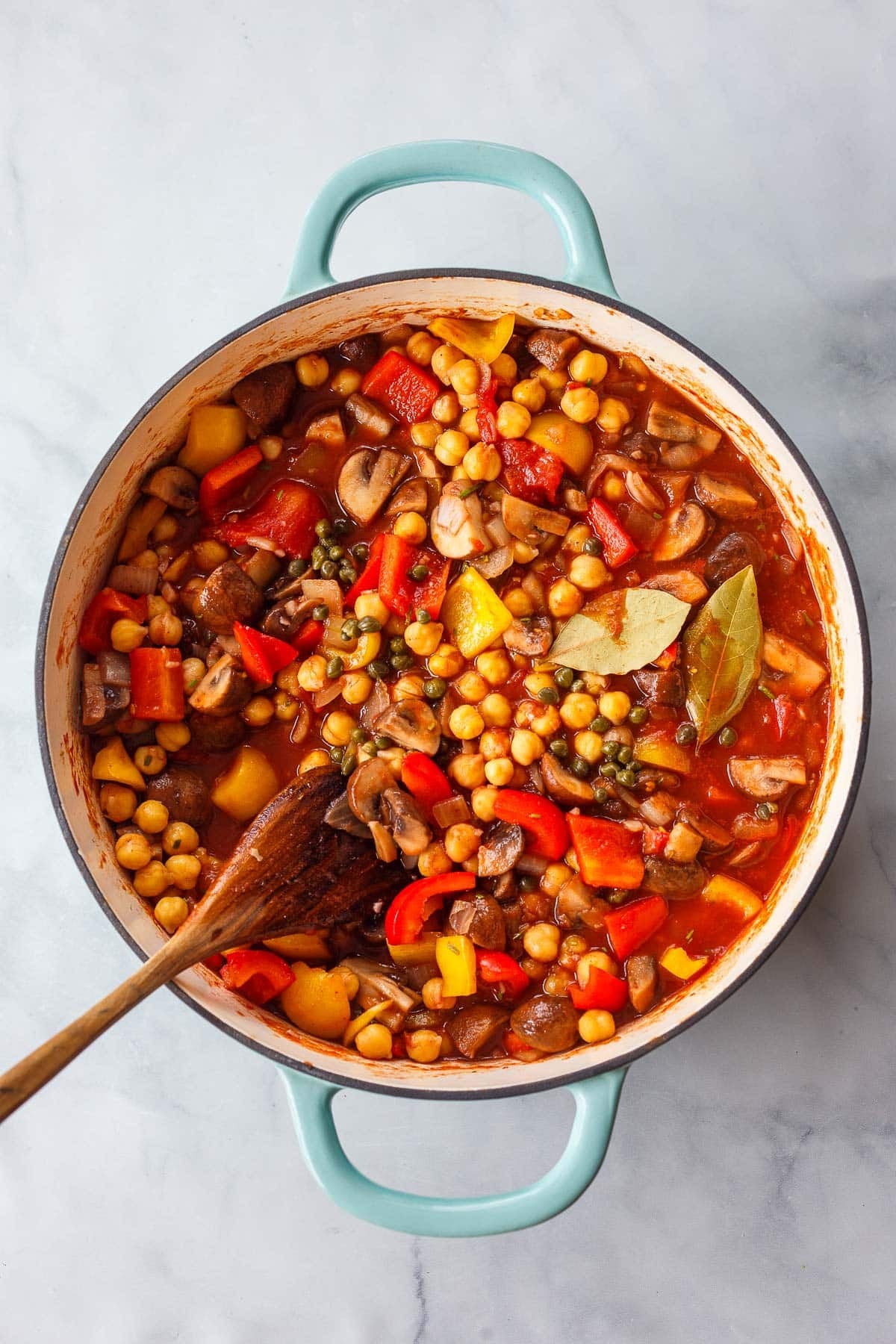 chickpea cacciatore in a dutch oven with peppers, mushrooms, capers, and bay leaves, with a wood spoon.