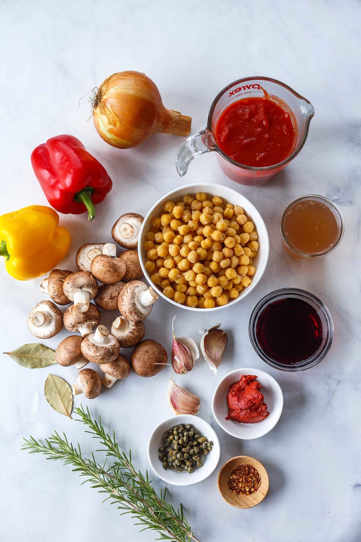 ingredients for chickpea cacciatore - onion, measuring cup of crushed tomatoes, red bell pepper, yellow bell pepper, bowl of chickpeas, jar of broth, mushrooms, bay leaves, garlic cloves, glass bowl of red wine, tomato paste in small white bowl, capers in small white bowl, fresh rosemary, and pinch bowl of red pepper flakes.