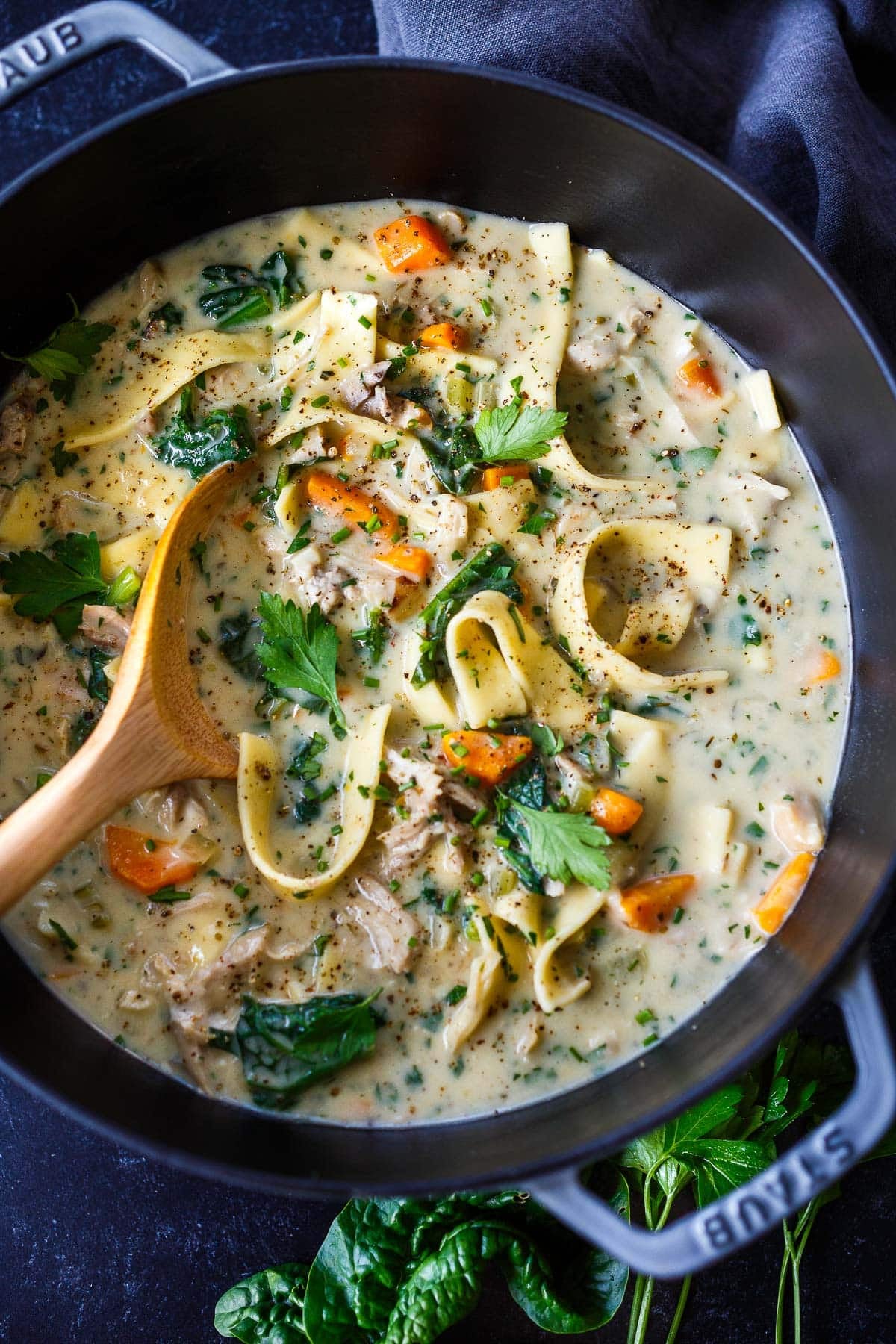 creamy chicken noodle soup in a dutch oven with a wooden spoon, with carrots, parsley, chives, spinach, and black pepper.