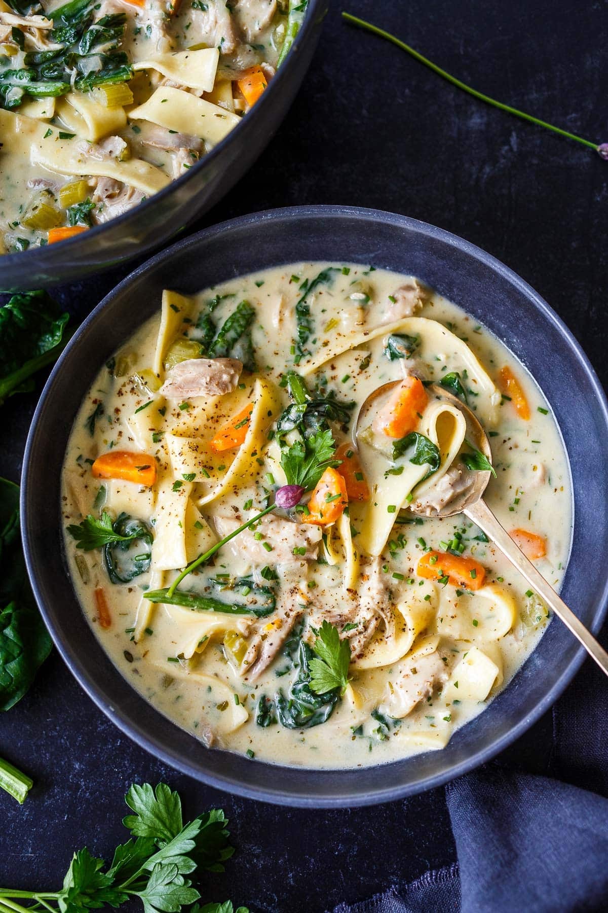 bowl of creamy chicken noodle soup with carrots, spinach, parsley, chives, and chili flakes, with a spoon.