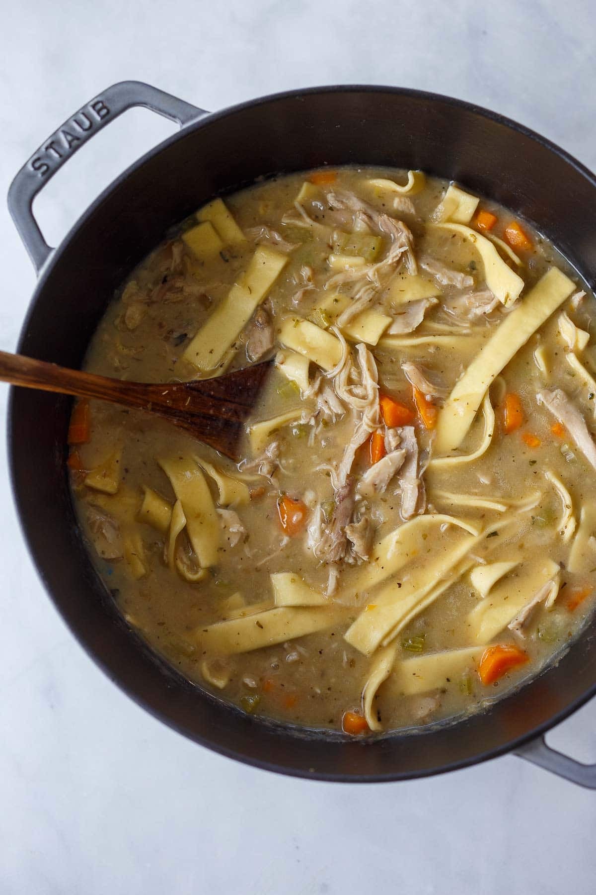 creamy chicken noodle soup cooking in a dutch oven with a wooden spoon.