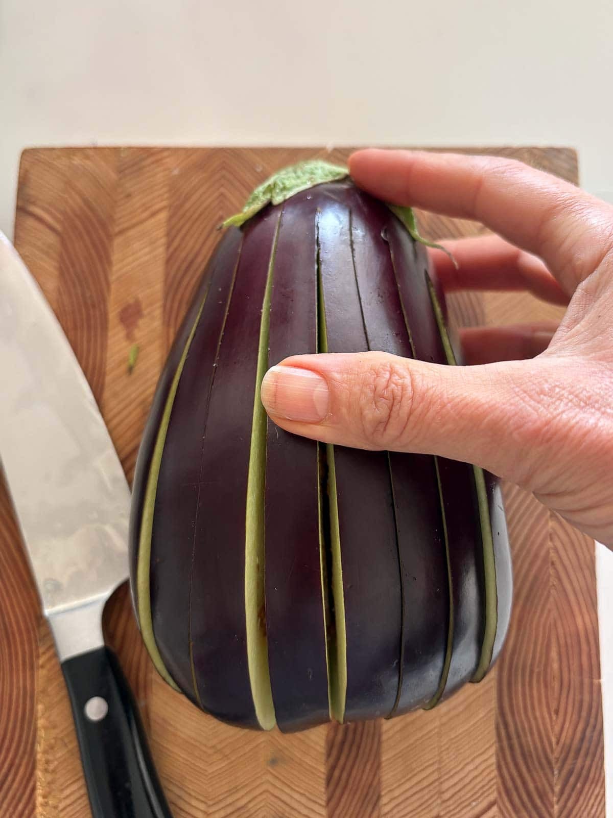 hand holding sliced eggplant with the stem intact on a wood cutting board next to a sharp knife.