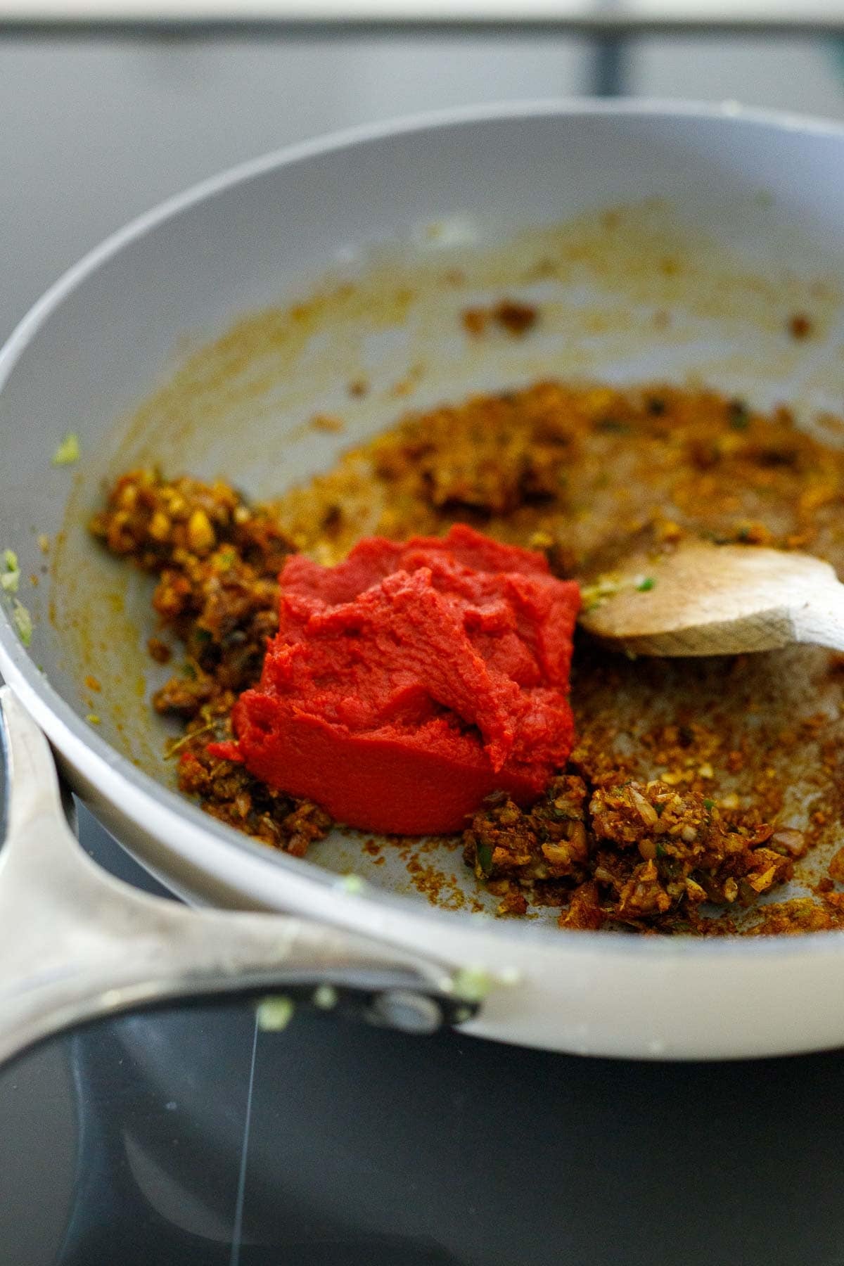 Indian spices, aromatics, and tomato paste in a skillet with a wooden spoon.