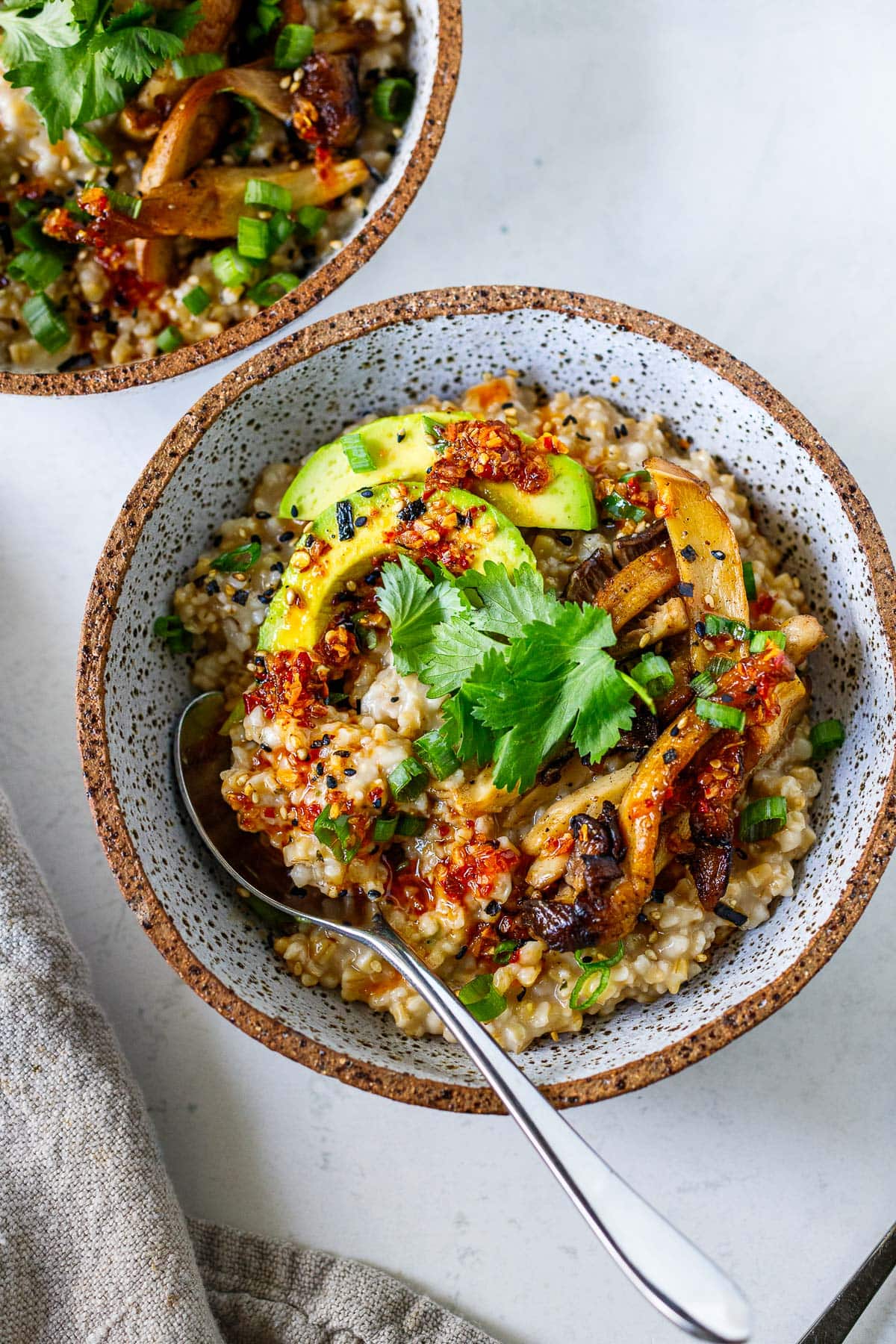 savory oats in a bowl with sliced avocado, sautéed mushrooms, cilantro leaves, sliced scallions, chili crisp, and furikake, with a spoon.