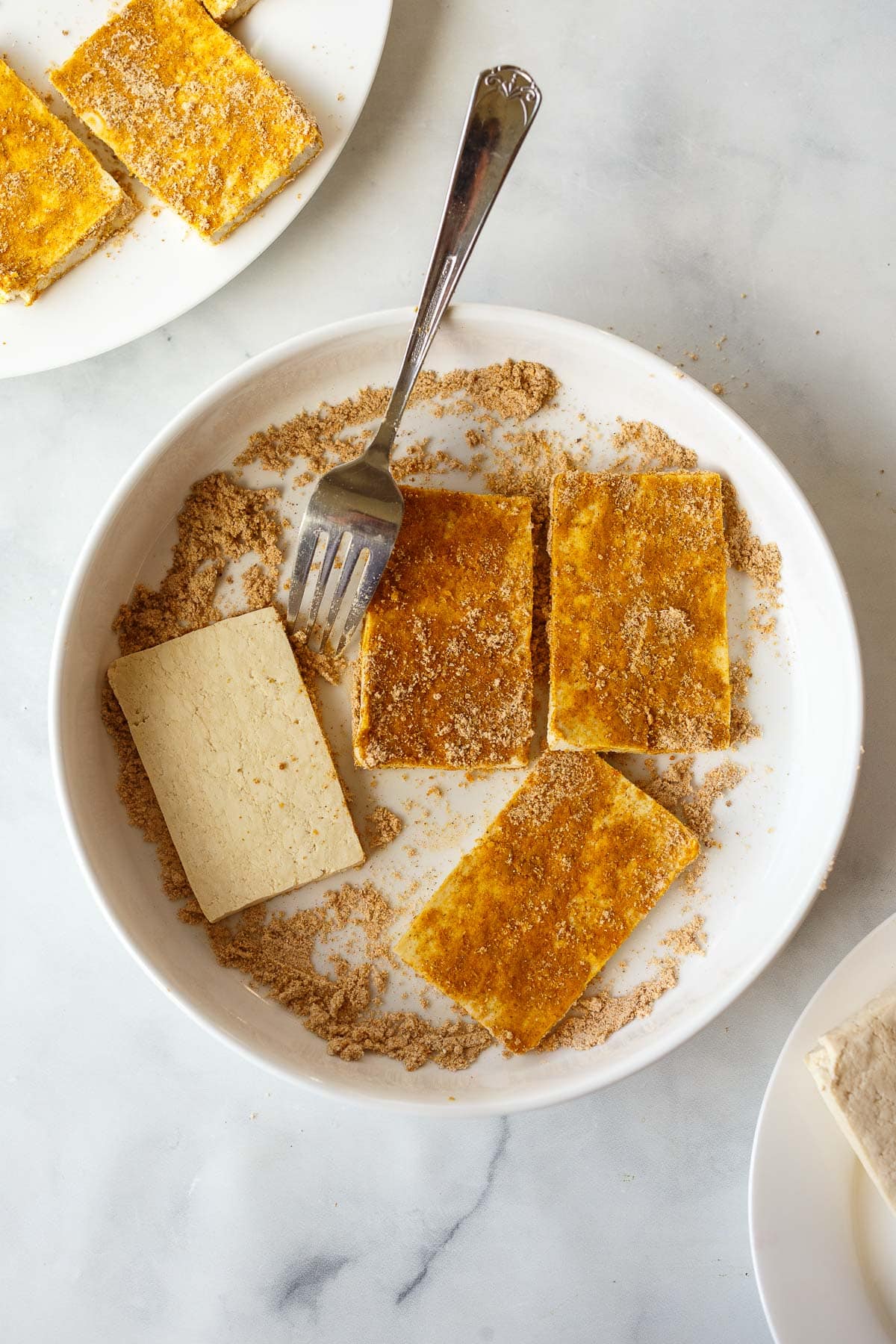 tofu slabs on a plate with shawarma spice blend and cornstarch, with a fork to flip the pieces.