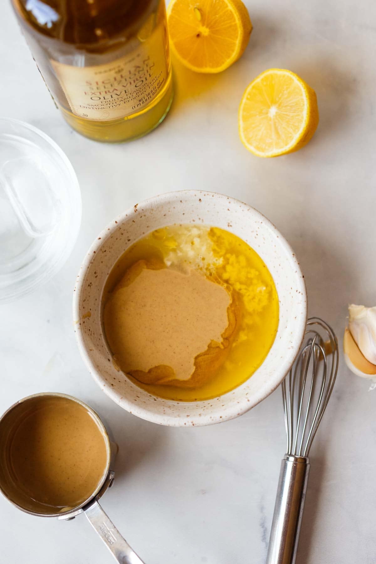 tahini sauce in a bowl next to lemons, olive oil, whisk, garlic cloves, and tahini sauce.