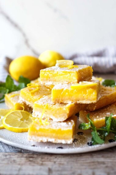 stack of four lemon bars on a plate with lemon slices and powdered sugar and mint leaves.
