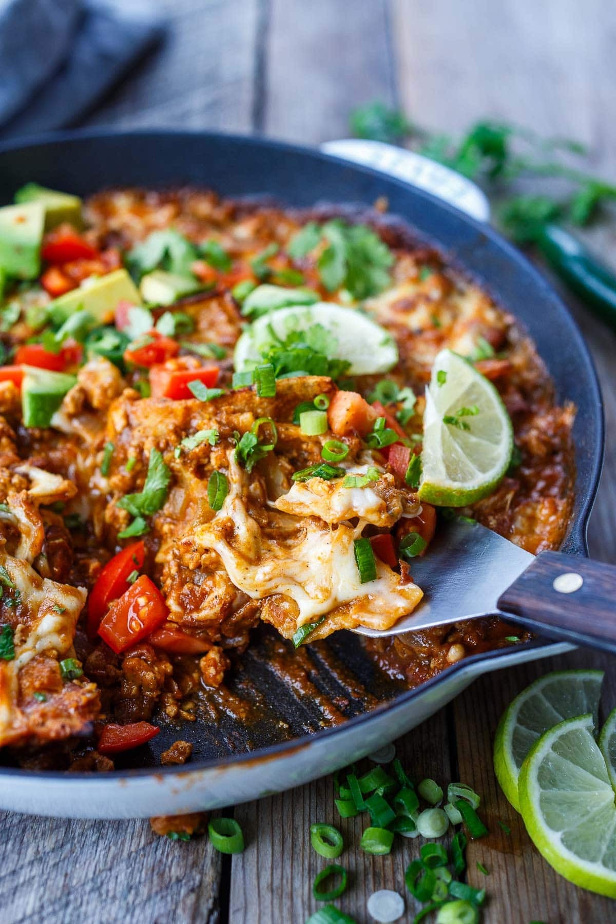 spatula scooping out a serving of enchilada skillet with lime wedges, scallions, fresh tomatoes, and herbs.