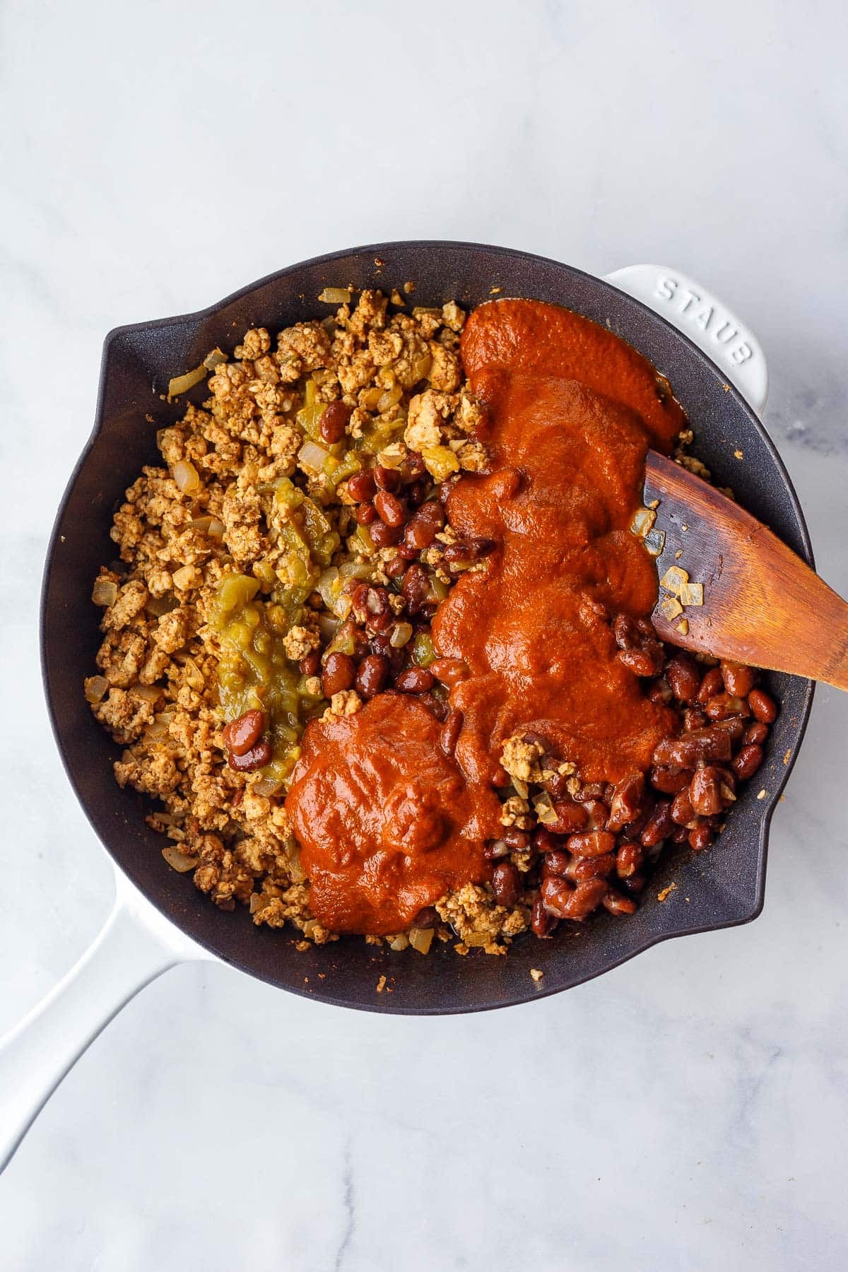 adding enchilada sauce, beans, and green chiles to a skillet of ground chicken.