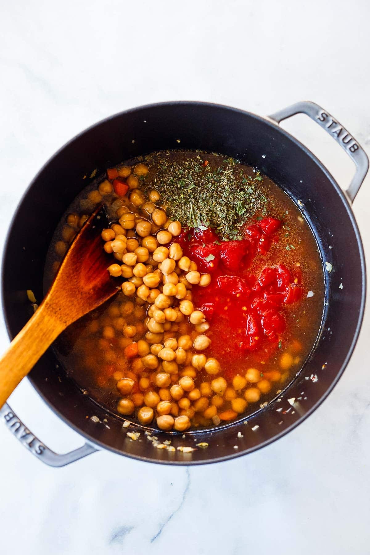 ingredients to make pasta e ceci in a pot with a wooden spatula, including chickpeas, tomatoes, and Italian seasoning. 