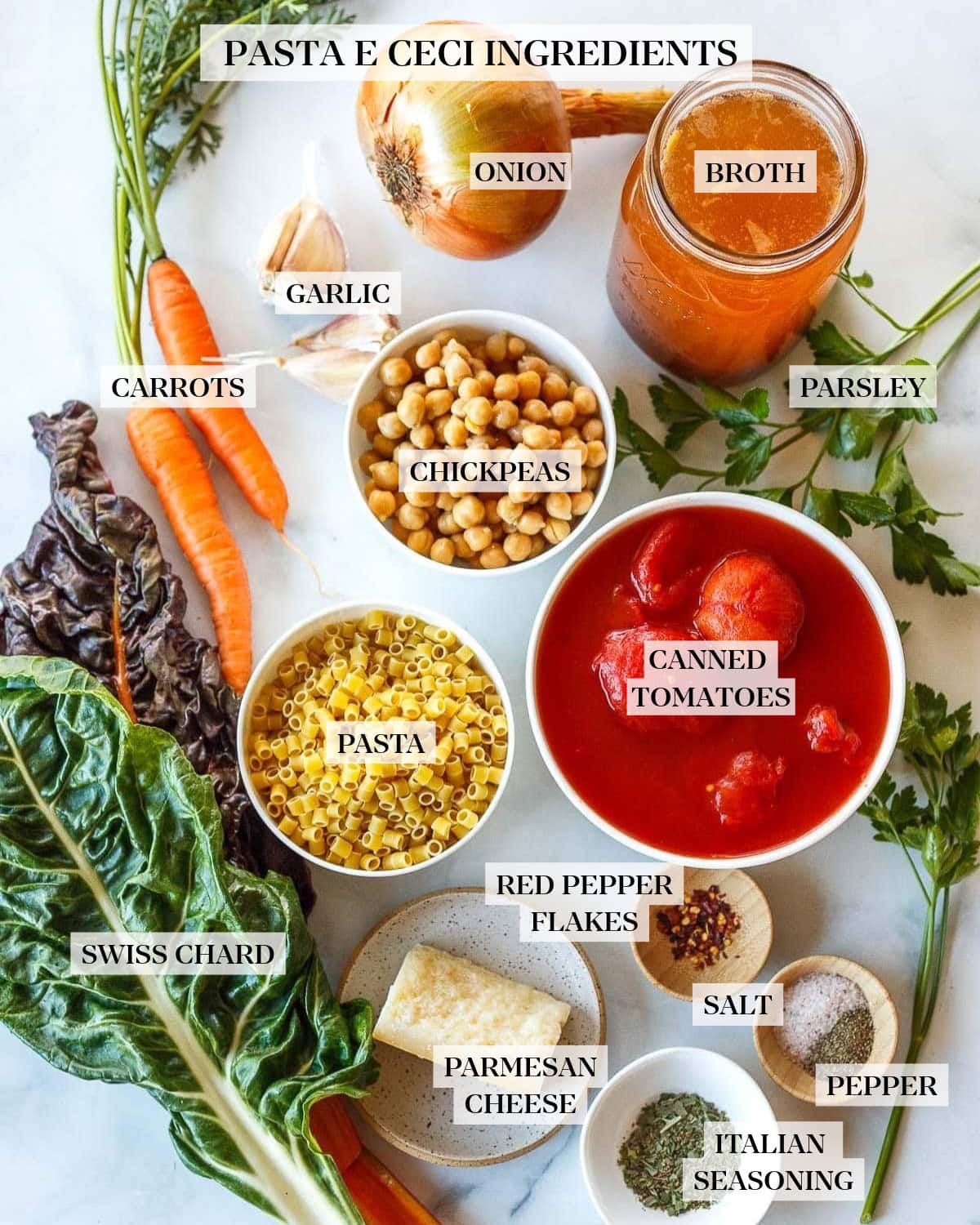 ingredients for pasta e ceci - onion, jar of broth, garlic cloves, carrots, bowl of chickpeas, ditalini pasta, and whole tomatoes, fresh parsley, chard, parmesan block, and pinch bowls of red pepper flakes, salt and pepper, and Italian seasoning. 
