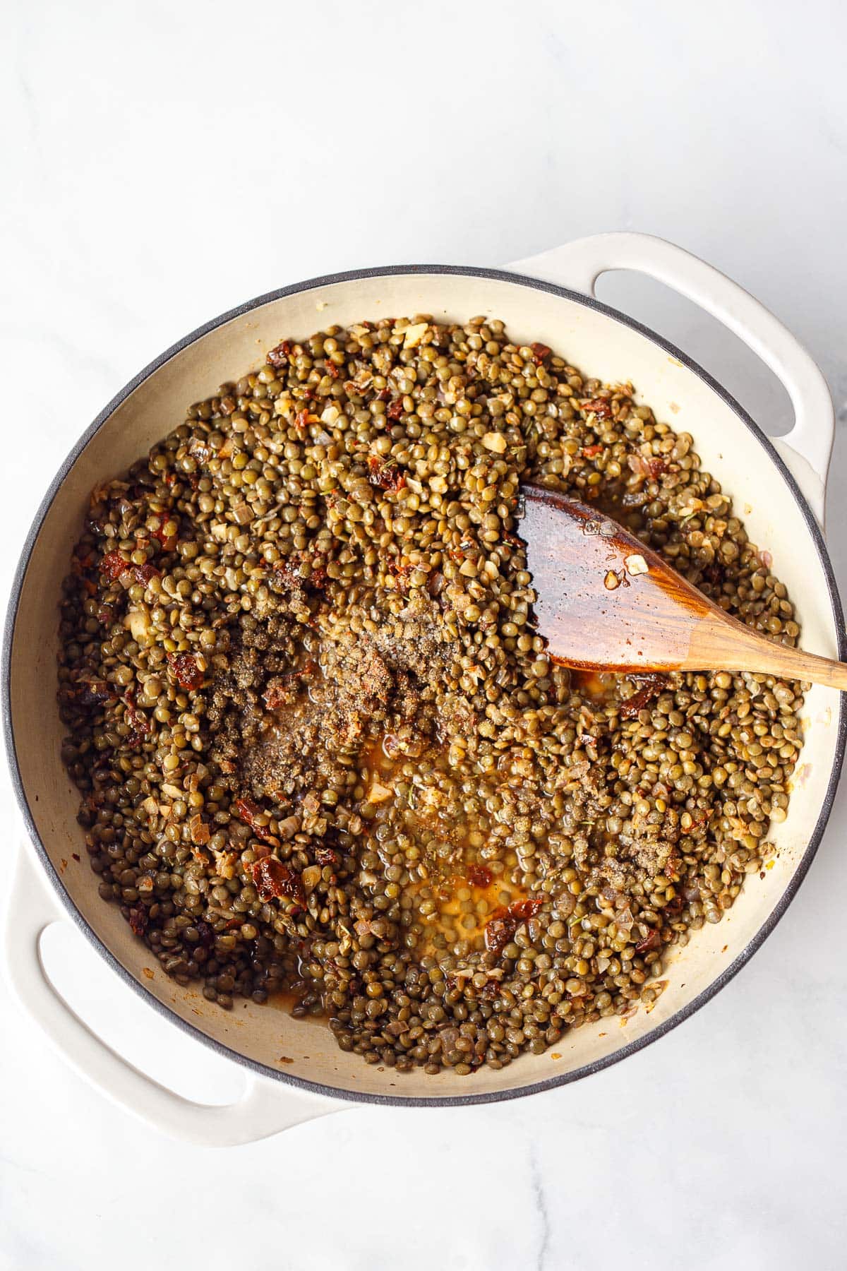 French green lentils in a pot with sun-dried tomatoes and broth, with a wooden spoon to stir. 