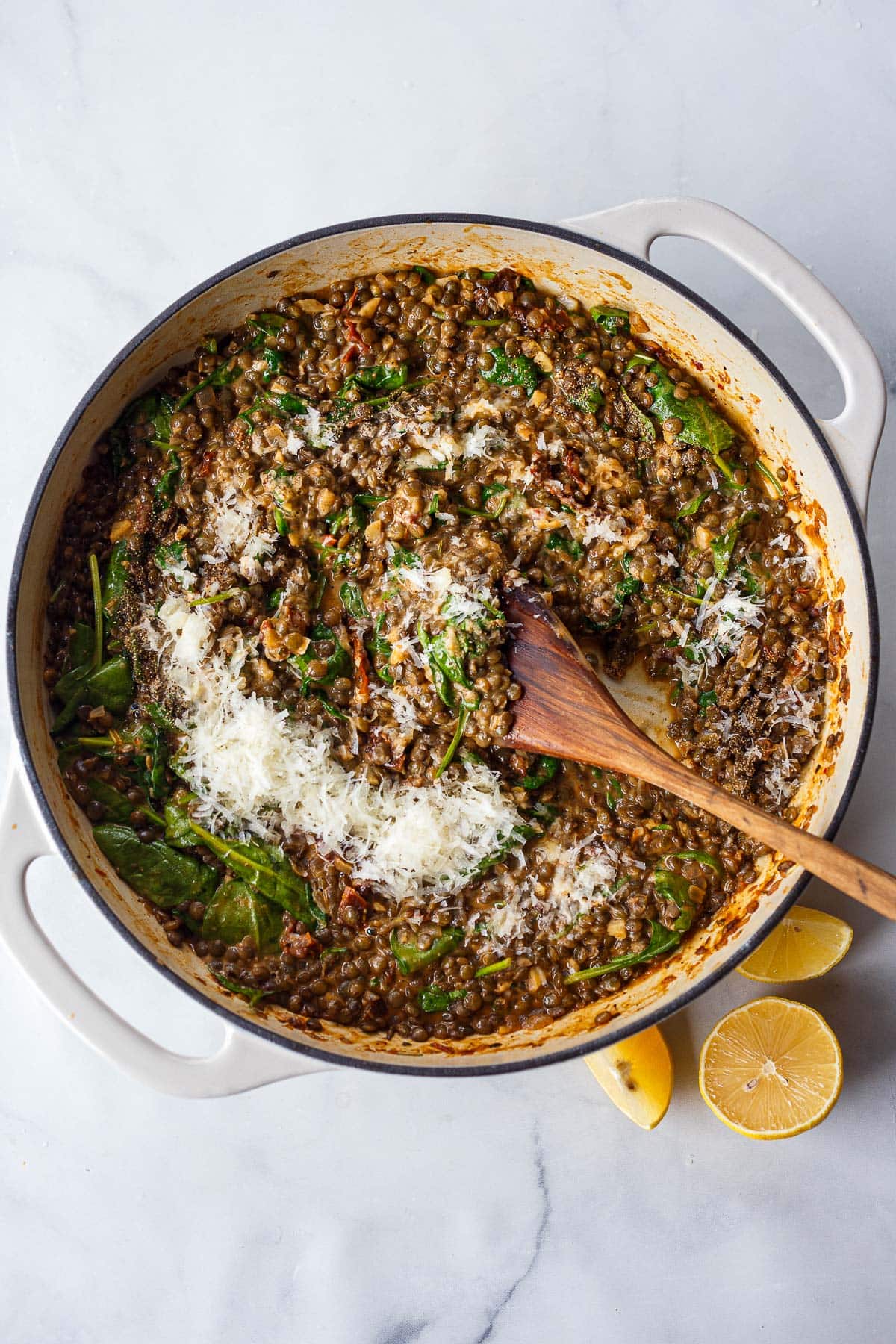 wood spoon stirring shaved Pecorino into a pot of Creamy Tuscan Lentils with lemon wedges on the side.