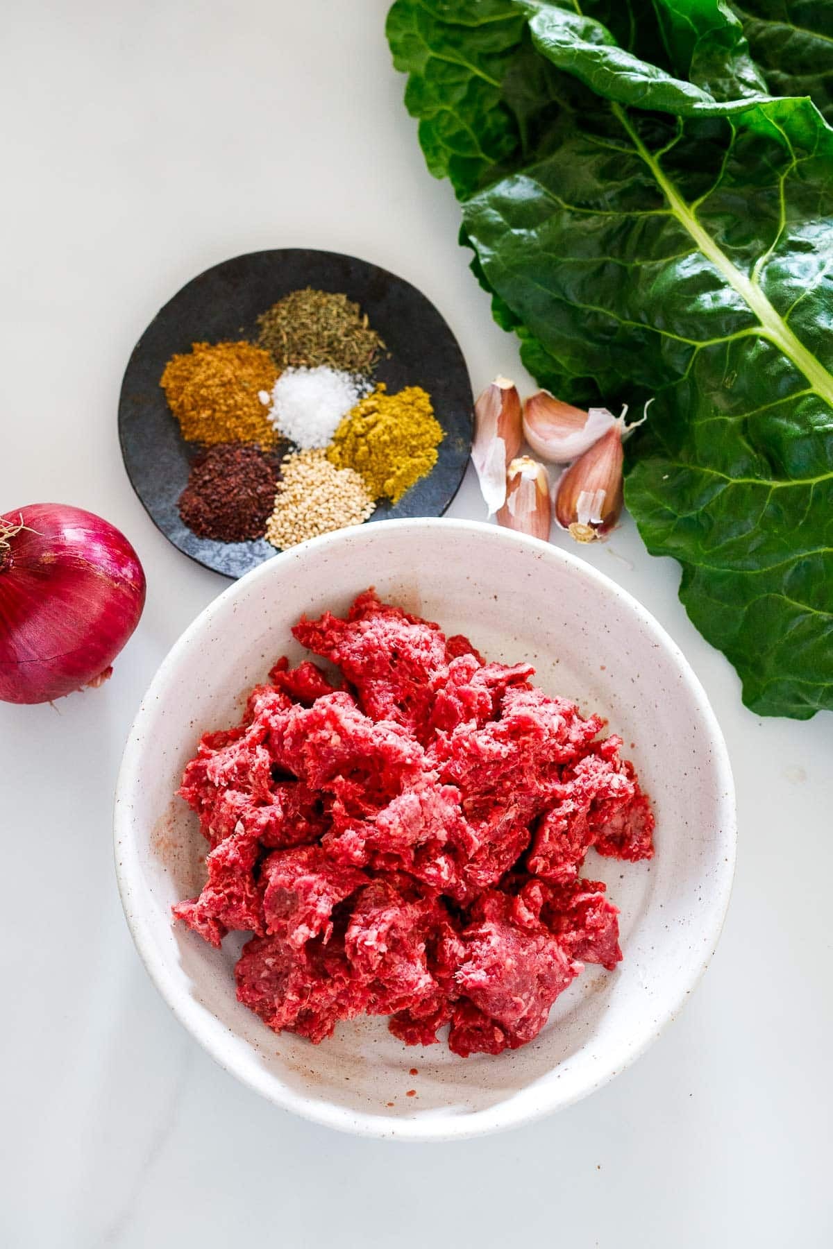 ingredients for zaatar beef and greens - chard, garlic cloves, zaatar spices, red onion, bowl of ground beef.