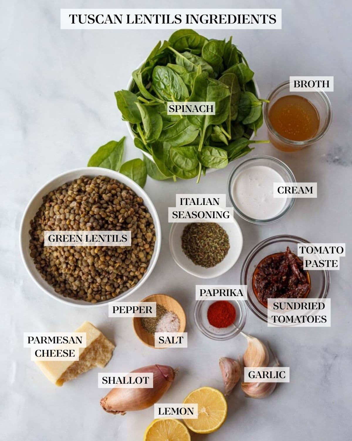 creamy Tuscan lentils ingredients neatly arranged on a marble countertop - bowl of spinach, glass bowls of broth, cream, cumin seeds, sun-dried tomatoes, smoked paprika, and a wood pinch bowl of salt and pepper, bowl of cooked French green lentils, Pecorino cheese, garlic cloves, shallot, and two lemon halves. 
